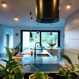 Artem Joanna Francuz-Płaza - Nowoczesna kuchnia z wyspą, czarnym zlewem i baterią, widok na jadalnię z dużym oknem i bukietem słoneczników, rośliny doniczkowe na pierwszym planie.