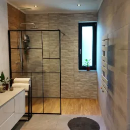 Nowoczesna łazienka z kabiną prysznicową typu walk-in z czarnymi szprosami, jasnymi płytkami imitującymi drewno na ścianach i podłodze, oraz białymi meblami łazienkowymi i grzejnikiem.