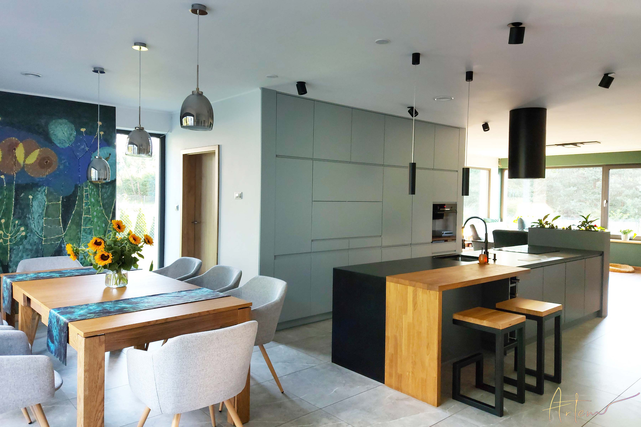 Nowoczesna kuchnia z wyspą, szarymi szafkami, drewnianym blatem i stołkami barowymi; na stole bukiet słoneczników i abstrakcyjny obraz na ścianie.