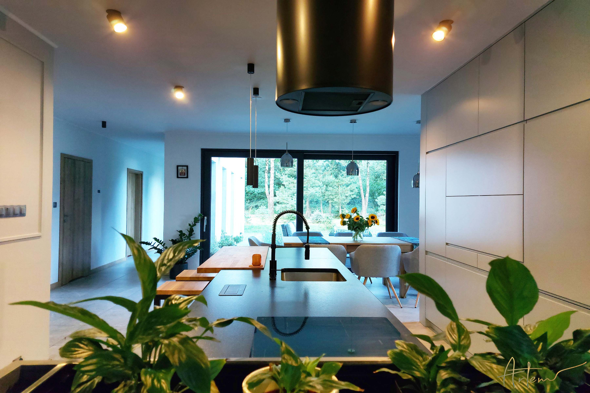 Nowoczesna kuchnia z wyspą, czarnym zlewem i baterią, widok na jadalnię z dużym oknem i bukietem słoneczników, rośliny doniczkowe na pierwszym planie.