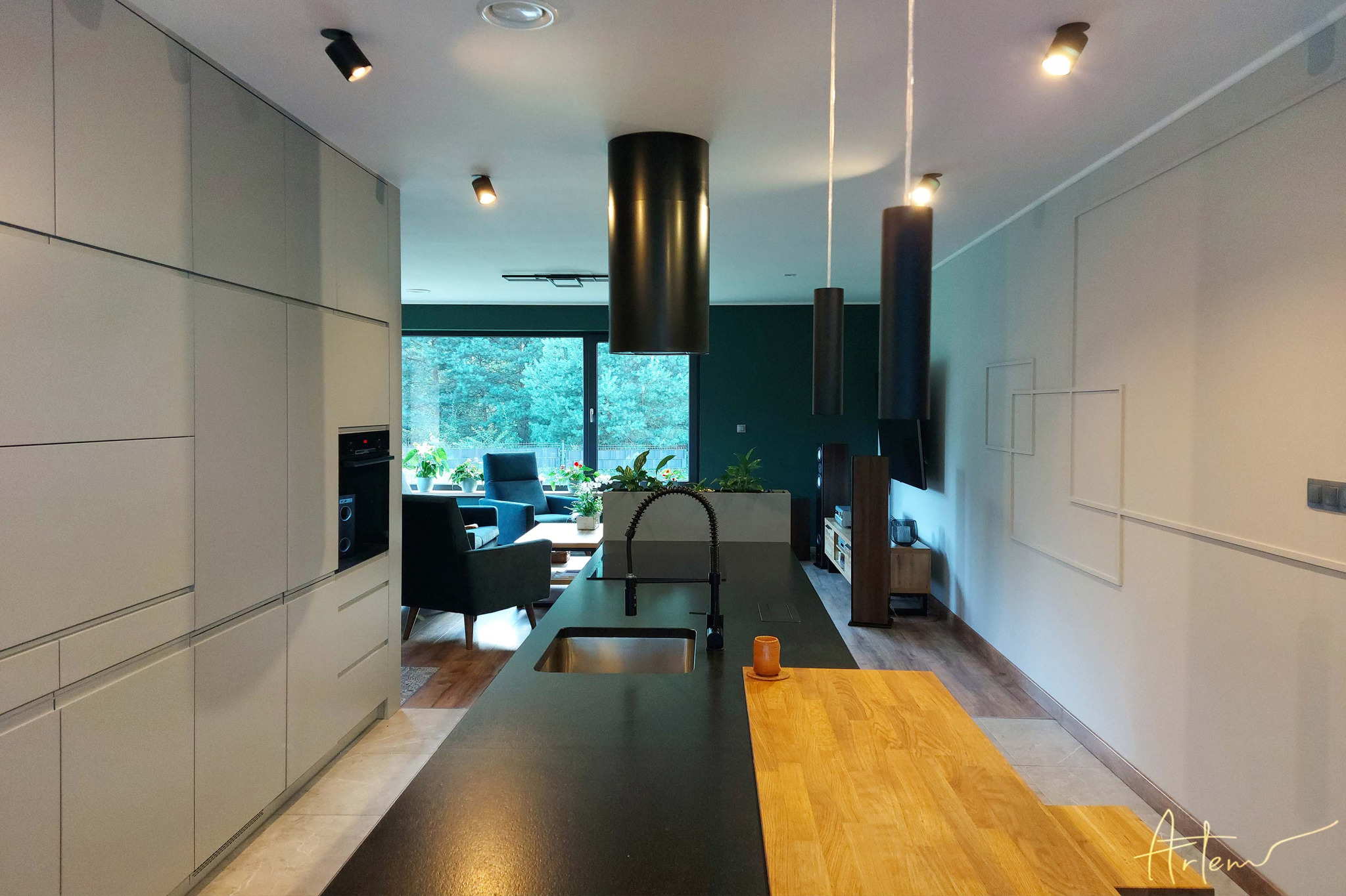 Nowoczesna kuchnia z wyspą, czarnym blatem i drewnianym elementem, widok na salon z zielonymi ścianami i roślinami w donicach, oświetlenie punktowe i wiszące lampy tuby.