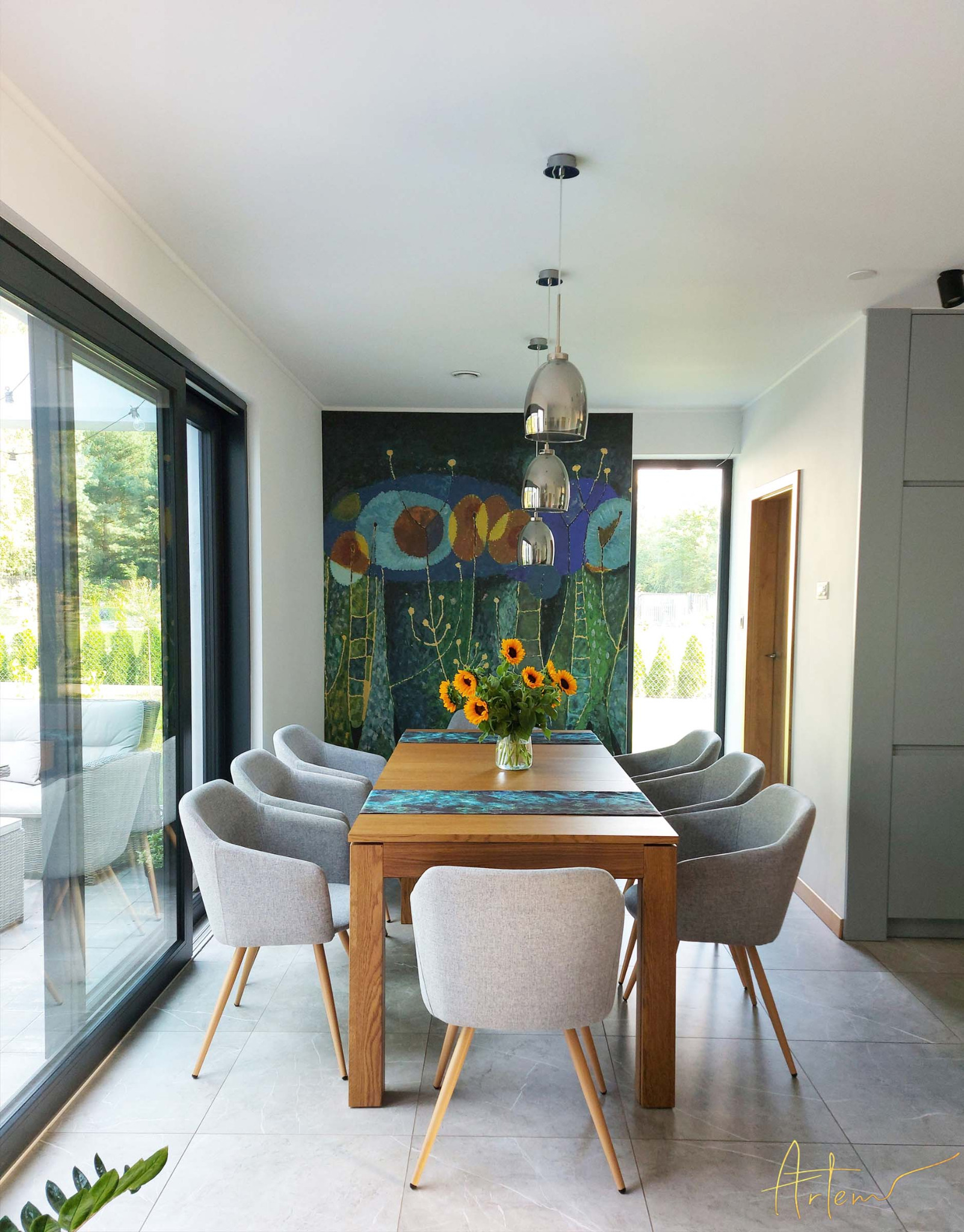 Jasne wnętrze jadalni z drewnianym stołem, szarymi krzesłami, bukietem słoneczników i abstrakcyjnym obrazem na ścianie, widok na taras przez przesuwne okno.