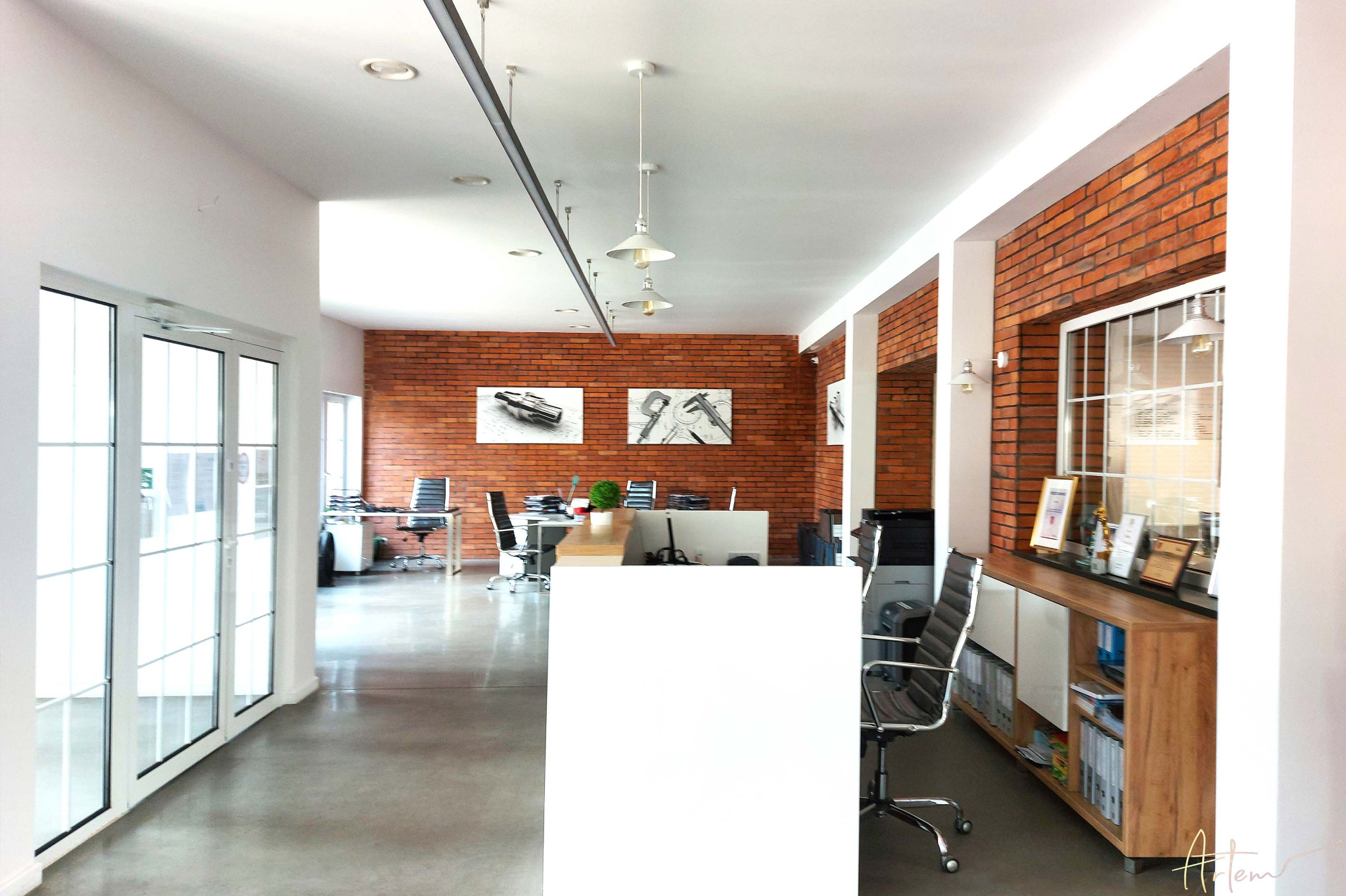 Nowoczesne biuro z ceglaną ścianą, białymi meblami i szarym betonem na podłodze, oświetlone wiszącymi lampami i ozdobione czarno-białymi grafikami technicznymi.
