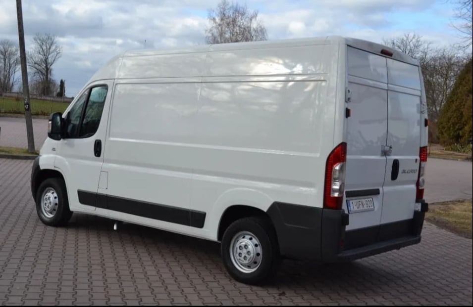 Biały bus dostawczy marki Fiat Ducato z belgijską tablicą rejestracyjną, zaparkowany na brukowanej powierzchni w pochmurny dzień.