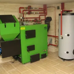 Zainstalowany, nowoczesny, zielony piec na paliwo stałe marki Kotrem z zasobnikiem, połączony z szarym zbiornikiem akumulacyjnym i czerwonymi rurami w kotłowni wykończonej płytkami imitującymi drewno.