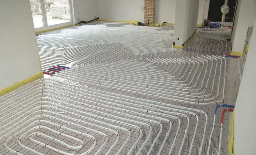 Rozłożone rury ogrzewania podłogowego na siatce, widoczne niebieskie i czerwone złączki, żółta taśma maskująca przy ścianach, surowy stan wykończenia wnętrza.