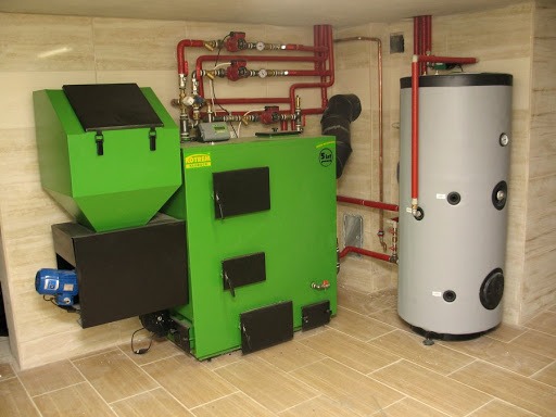Zainstalowany, nowoczesny, zielony piec na paliwo stałe marki Kotrem z zasobnikiem, połączony z szarym zbiornikiem akumulacyjnym i czerwonymi rurami w kotłowni wykończonej płytkami imitującymi drewno.