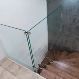 Widok z góry na spiralne schody z drewnianymi stopniami i przezroczystą szklaną balustradą mocowaną za pomocą metalowych uchwytów, na tle białej ściany i szarej ściany o fakturze betonu.