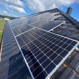 Panele słoneczne na dachu z ciemnej dach&oacute;wki, widok z dołu, niebieskie niebo z chmurami w tle, basen ogrodowy w oddali.