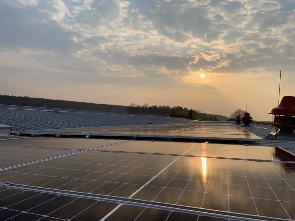 Instalacja paneli słonecznych na dachu przemysłowym, widok na zachodzące słońce odbijające się w panelach, w tle las i komin wentylacyjny.