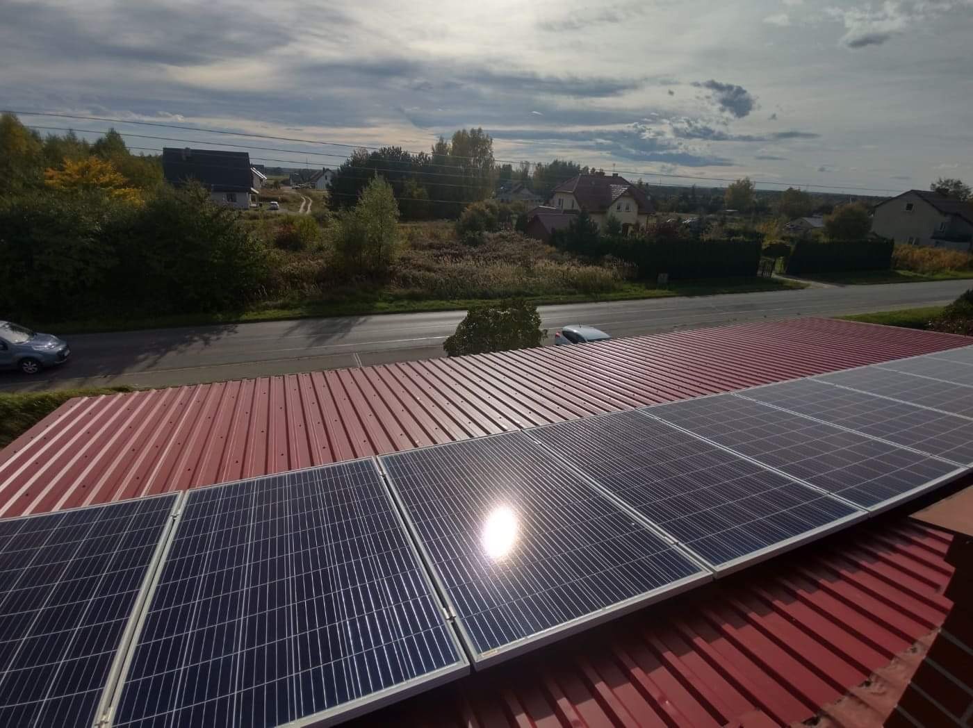 Panele słoneczne na czerwonym dachu blachodachówki, odbijające światło słoneczne, z widokiem na okoliczną zabudowę i drogę.