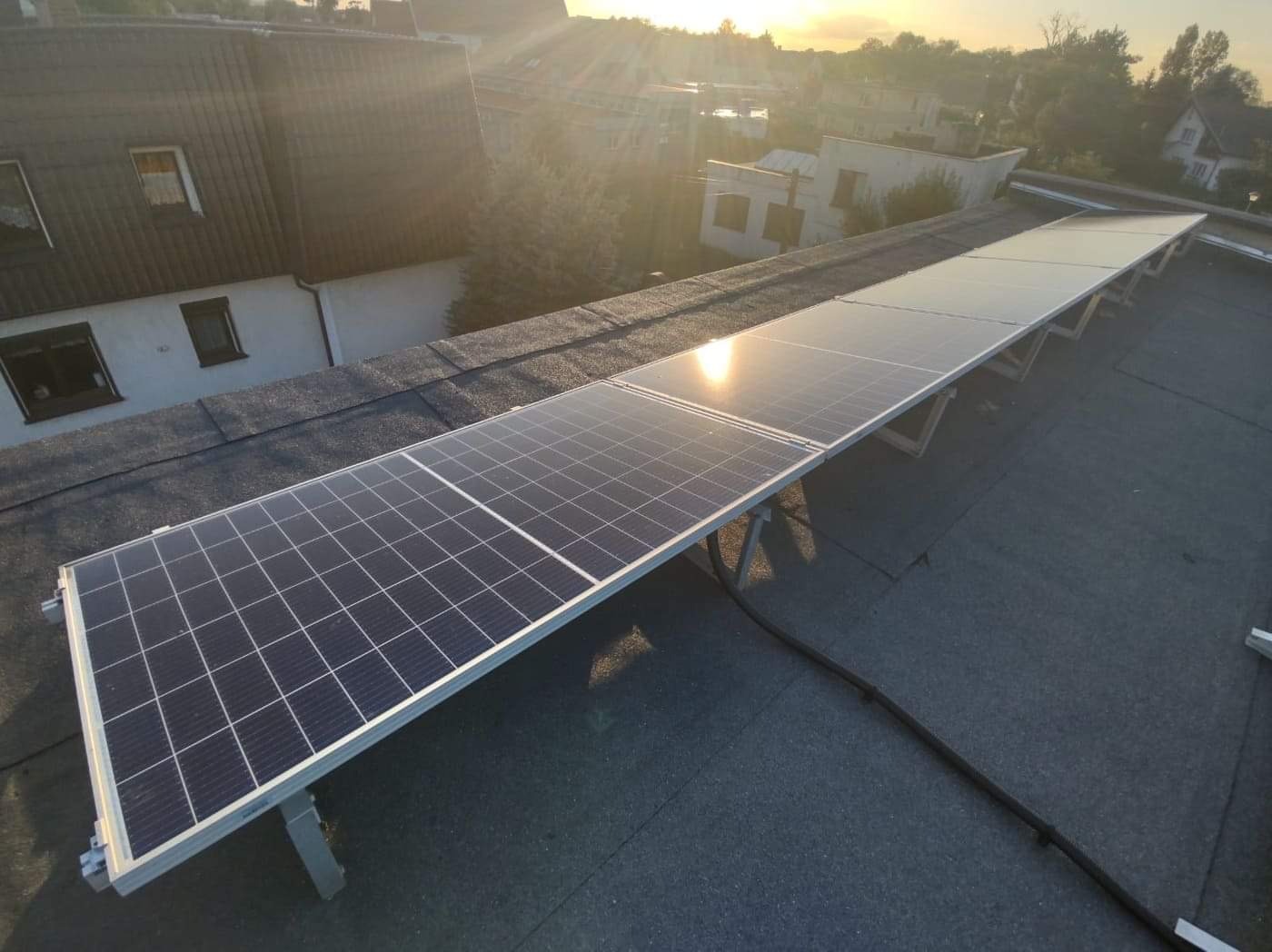 Rząd paneli słonecznych na płaskim dachu pokrytym papą w promieniach wschodzącego słońca, widok na zabudowania w tle.
