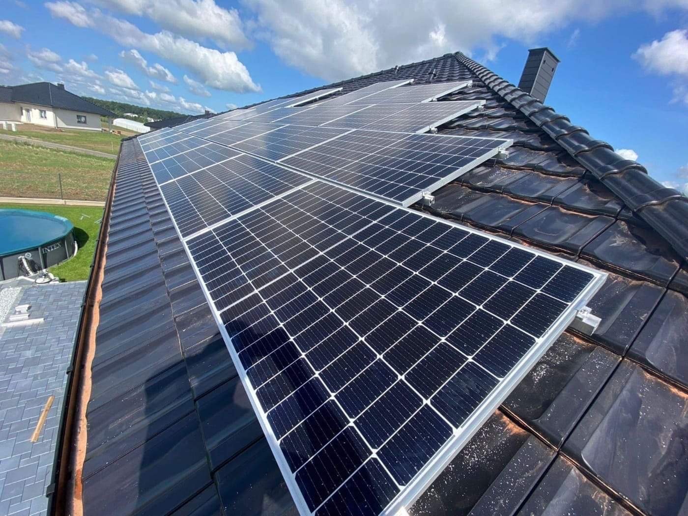 Panele słoneczne na dachu z ciemnej dachówki, widok z dołu, niebieskie niebo z chmurami w tle, basen ogrodowy w oddali.