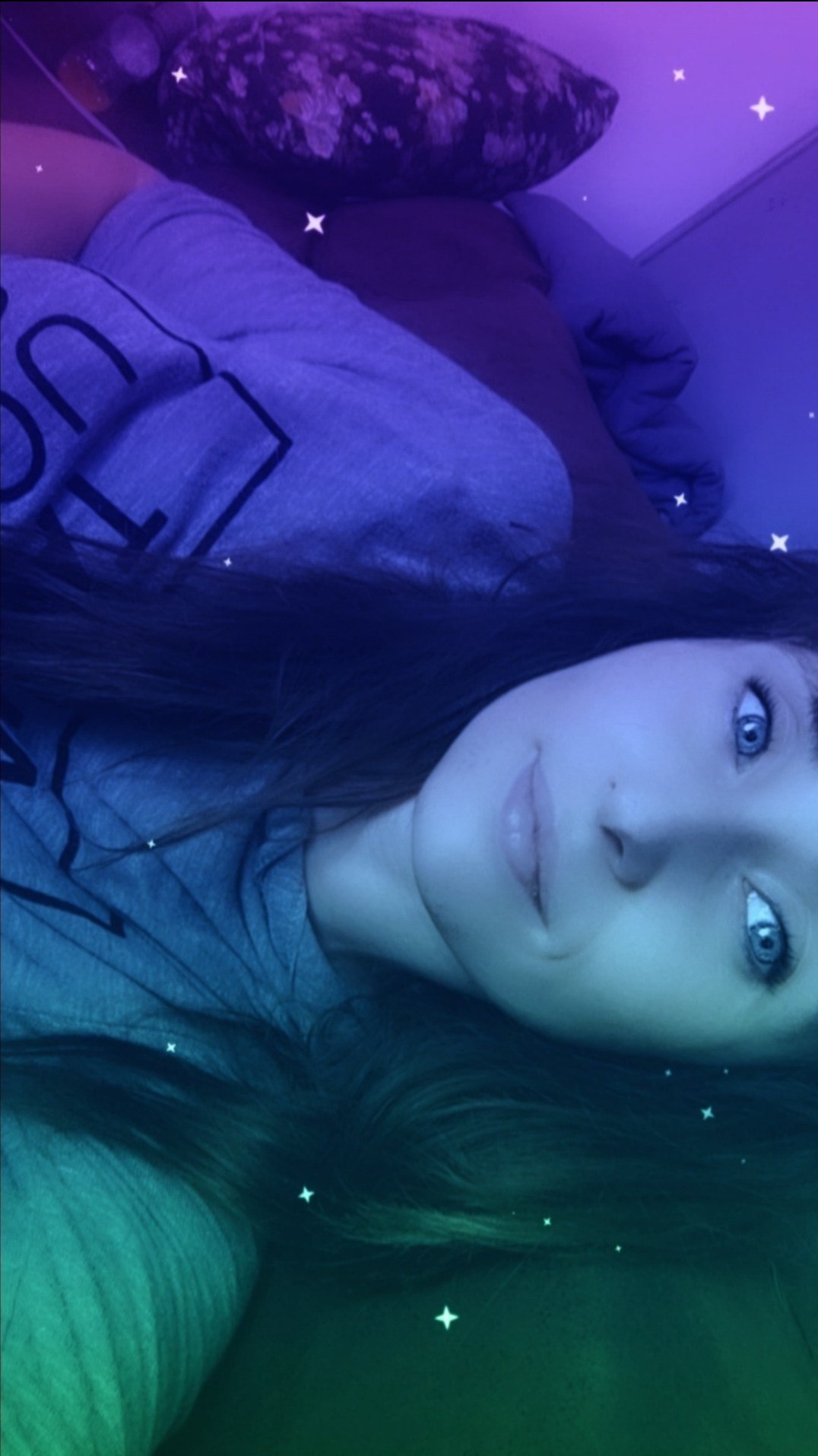 Portret młodej kobiety z długimi, ciemnymi włosami i niebieskimi oczami, leżącej na łóżku z wzorzystą poduszką, oświetlony niebiesko-zielonym filtrem i efektami gwiazd.