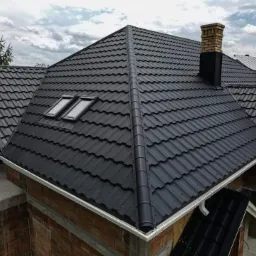 Czarny dach z blachodachówki z dwoma oknami dachowymi i ceglanym kominem, widoczny montaż rynny. Budynek w trakcie budowy.