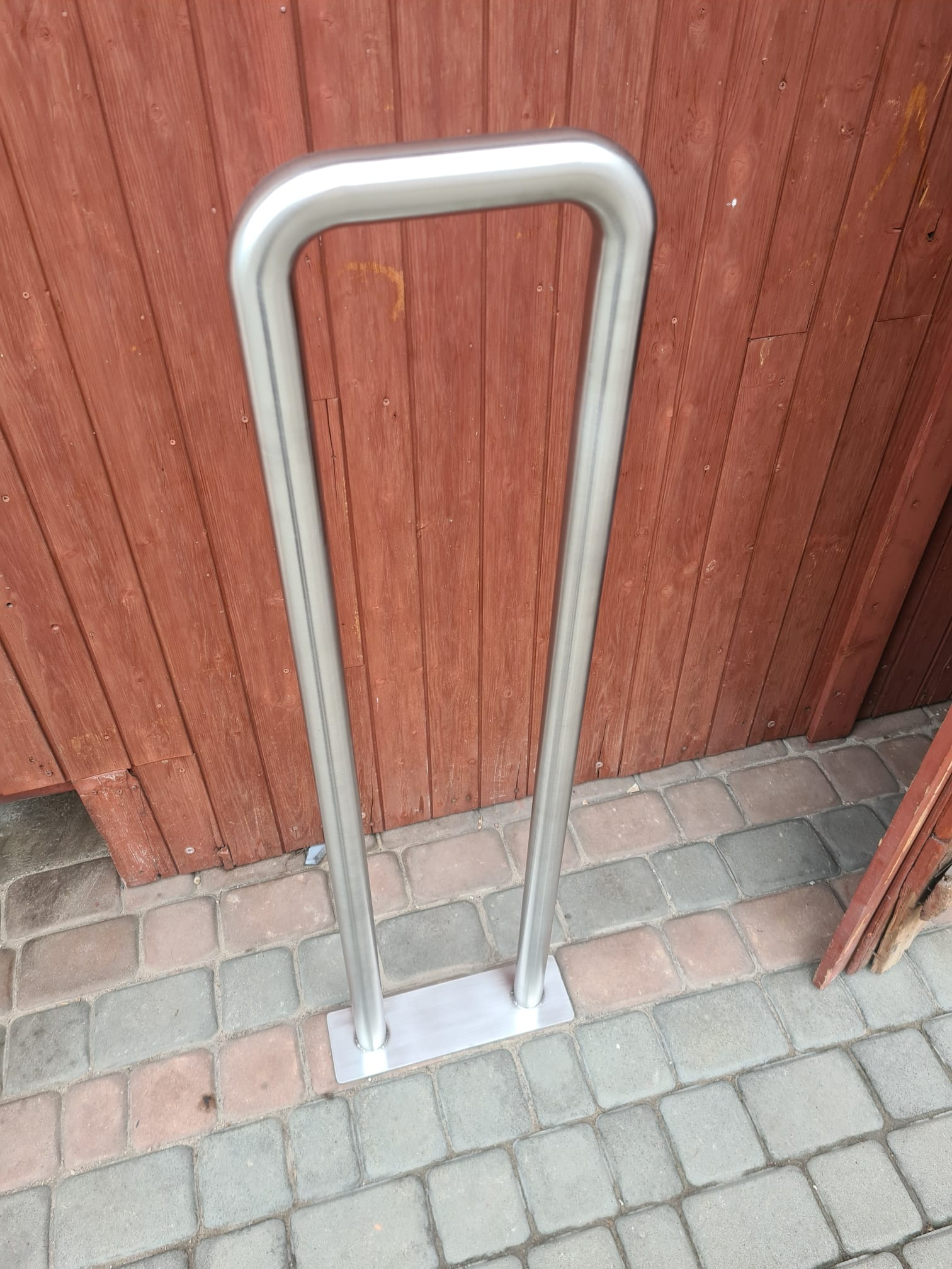 Stojak na rowery ze stali nierdzewnej, zamontowany na kostce brukowej przed drewnianą ścianą.