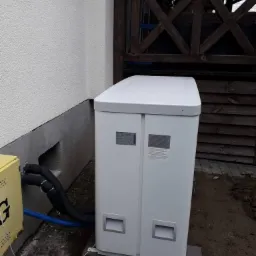 Biała pompa ciepła zamontowana na zewnątrz budynku, z widocznymi rurami przyłączeniowymi i fragmentem elewacji oraz drewnianego ogrodzenia.