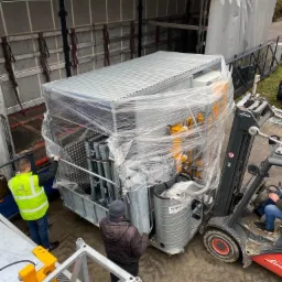 Załadunek metalowej konstrukcji windy towarowej, owiniętej folią, do naczepy ciężarówki za pomocą wózka widłowego Linde, widoczni pracownicy w odblaskowych kamizelkach.