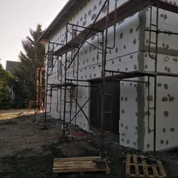 Dom w trakcie ocieplania, widoczne płyty styropianowe z zaprawą, rusztowanie metalowe, palety na ziemi i drzewa w tle.