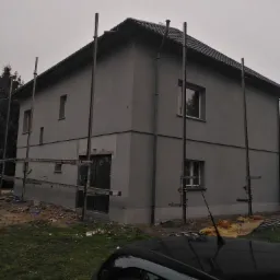 Dwukondygnacyjny budynek z szarą elewacją w trakcie prac wykończeniowych, widoczne rusztowania i resztki materiałów budowlanych na trawniku, fragment czarnego samochodu w dolnej części kadru.