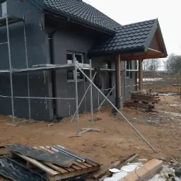 Dom jednorodzinny w trakcie ocieplania elewacji zewnętrznej, widoczne rusztowania i styropian grafitowy, dach pokryty blachodachówką w kolorze grafitowym, teren wokół domu niezagospodarowany.