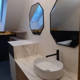 Nowoczesna łazienka na poddaszu ze skośnym sufitem, owalna biała umywalka nablatowa z czarną armaturą, dwa ośmiokątne lustra w czarnych ramach, marmurowy blat i okładzina ściany, widoczne okno...