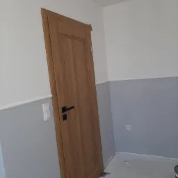 Świeżo pomalowane wnętrze z drewnianymi drzwiami z czarną klamką, biało-szare ściany z listwami dekoracyjnymi, widoczne puszki z farbą i wałek malarski na podłodze.