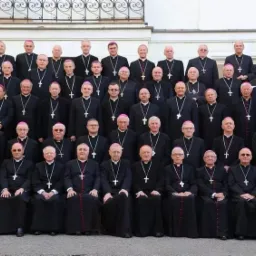 Grupowe zdjęcie biskupów w czarnych sutannach z krzyżami na szyjach, ustawionych w rzędach na tle jasnej ściany budynku.