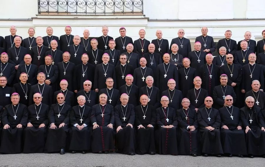 Grupowe zdjęcie biskupów w czarnych sutannach z krzyżami na szyjach, ustawionych w rzędach na tle jasnej ściany budynku.