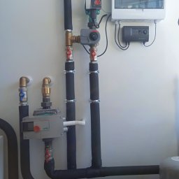 DE Systemy sp. z o. o. - Węzeł cieplny z rurami w izolacji, zaworami, pompą obiegową Wilo, skrzynką elektryczną i automatyką na białej ścianie.