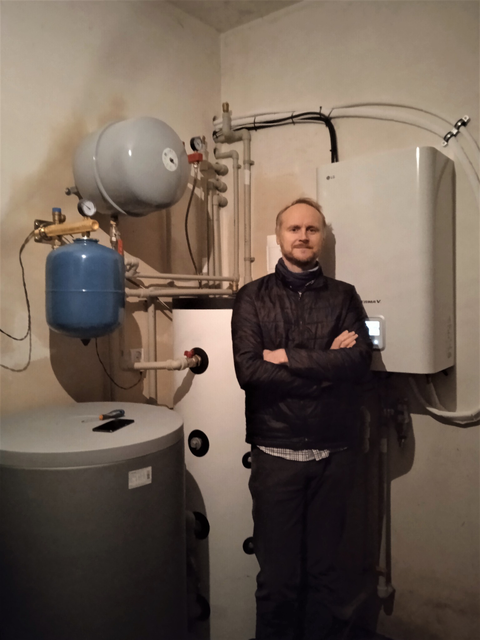 Mężczyzna z założonymi rękami stoi w kotłowni obok zbiornika buforowego, pompy ciepła LG Therma V i innych elementów instalacji grzewczej.