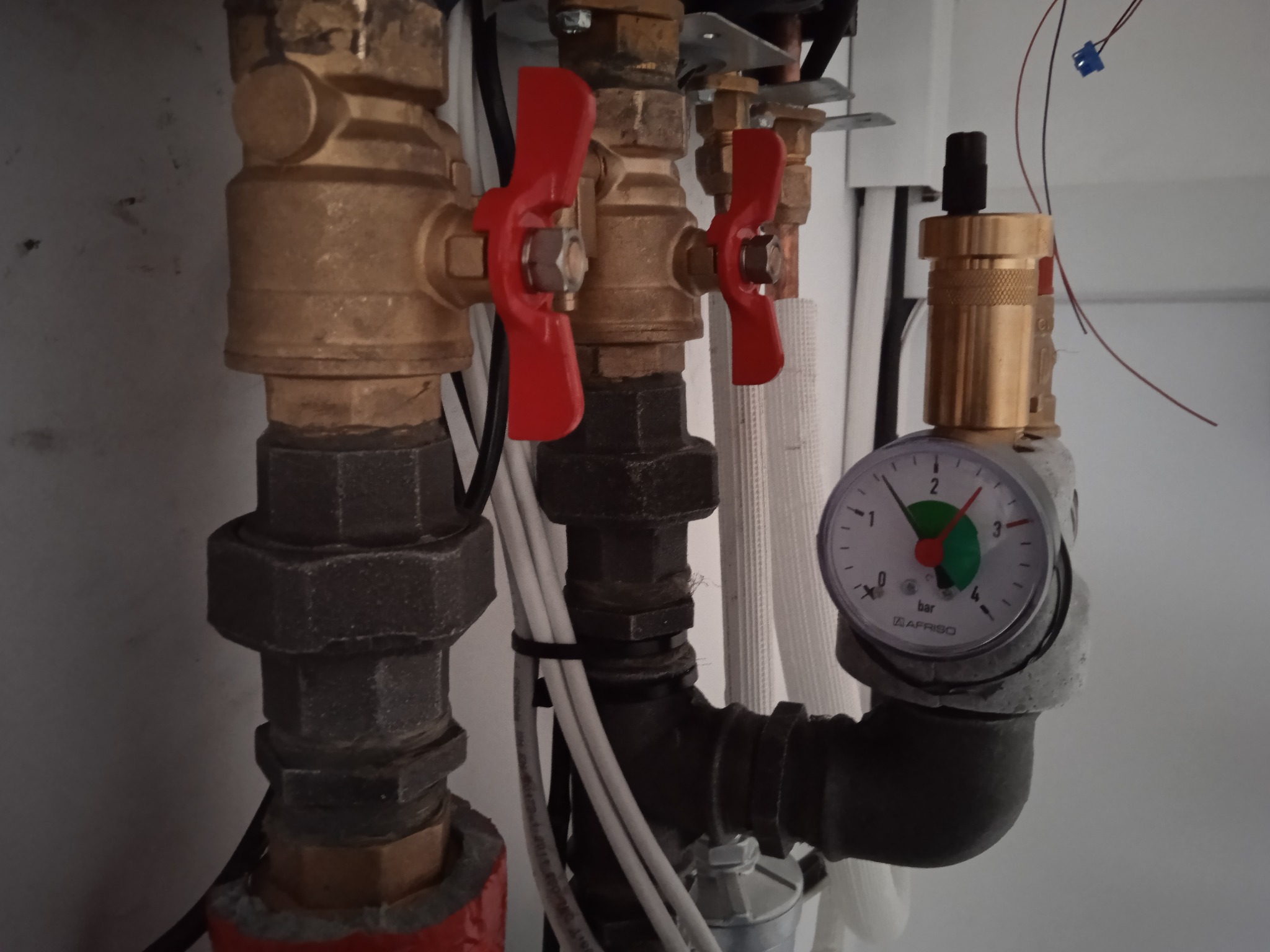Fragment instalacji z zaworami kulowymi z czerwonymi uchwytami i manometrem wskazującym ciśnienie w systemie, widoczne rury miedziane i elementy mocujące.