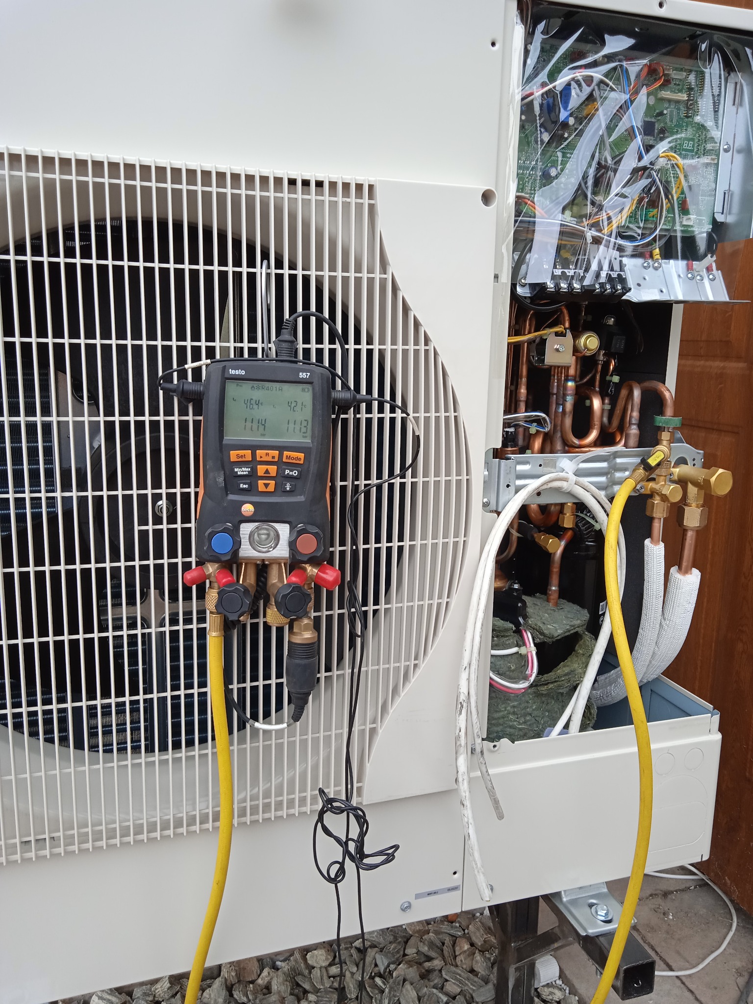Podłączenie cyfrowego manometru Testo 557 do pompy ciepła podczas serwisu, widoczne przewody pomiarowe, otwarty panel z elektroniką i rurami miedzianymi.