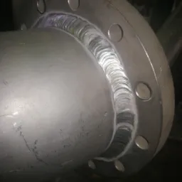 Szczegółowe zbliżenie spawu kołnierza do rury ze stali nierdzewnej, widoczne regularne ślady spawania TIG, zarysowania na powierzchni metalu.