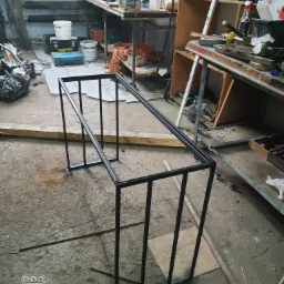 Surowa, czarna, metalowa rama stolika kawowego w warsztacie, widoczne ślady spawania, w tle narzędzia i materiały.