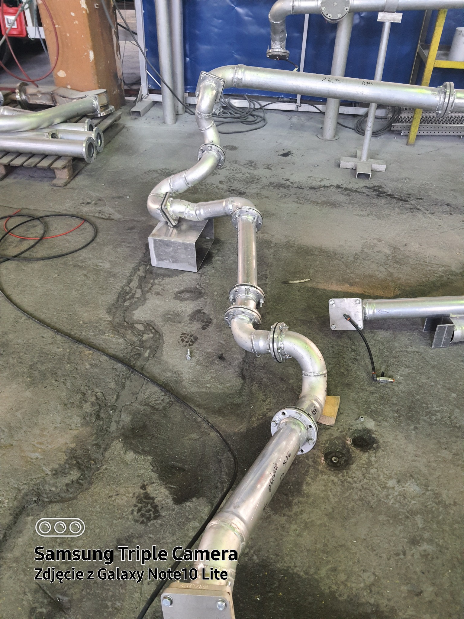 Elementy instalacji przemysłowej ze stali nierdzewnej, rury łączone kołnierzami, ułożone na szarym, betonowym podłożu w hali produkcyjnej, widoczne spawane łączenia i podpory.