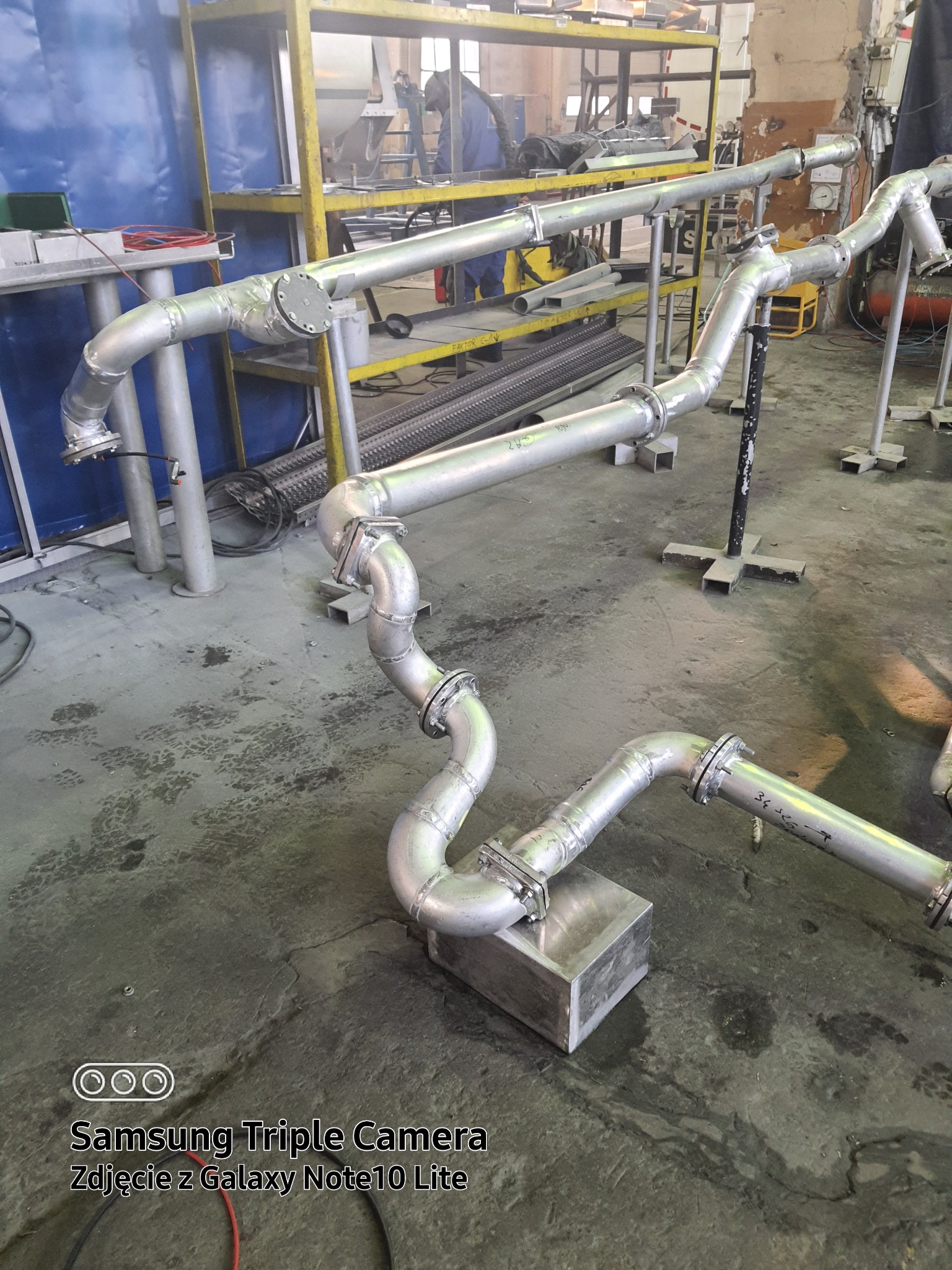 Złożony system rur ze stali nierdzewnej, z licznymi złączami kołnierzowymi i spawami, ustawiony w warsztacie z widocznym regałem i elementami konstrukcyjnymi w tle.