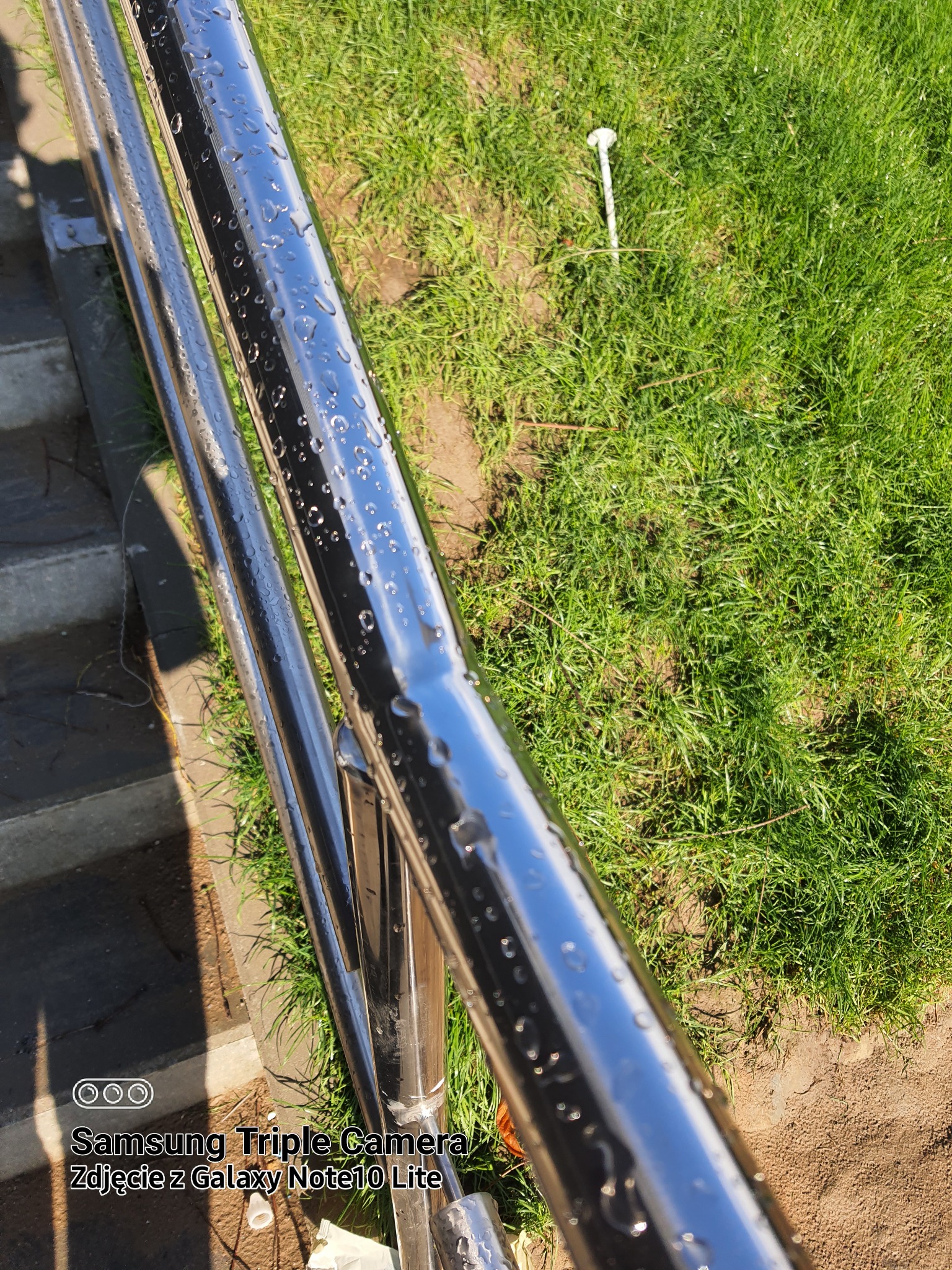 Błyszcząca, nierdzewna poręcz schodowa w Elblągu, pokryta kroplami wody, sfotografowana pod kątem, z widocznym trawnikiem w tle.