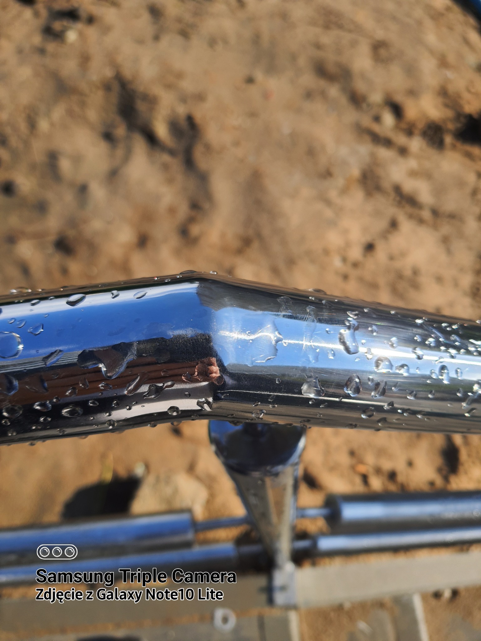 Błyszcząca poręcz nierdzewna z kroplami wody, widoczne odbicie otoczenia i fragment tła z ziemią.