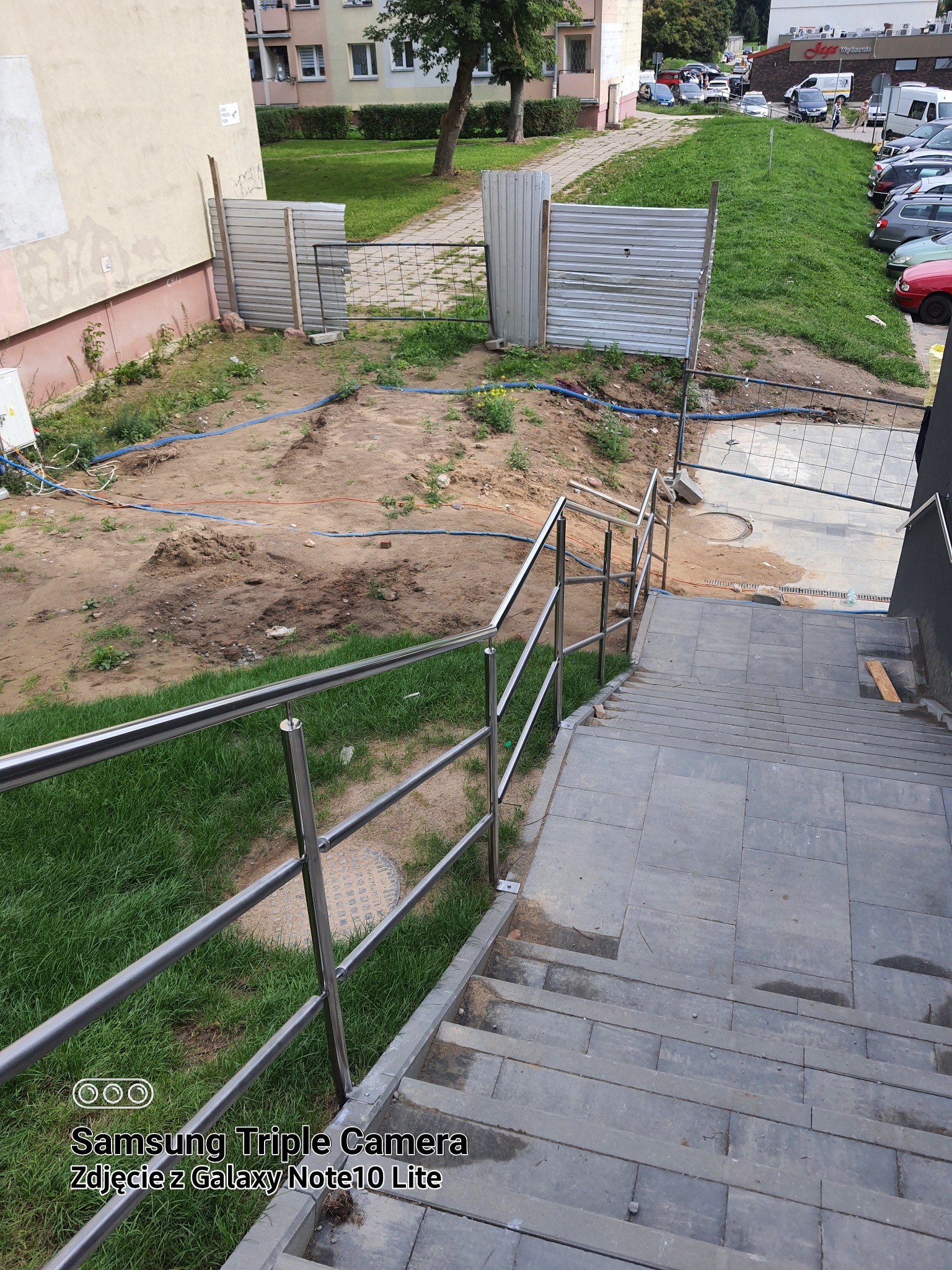 Zewnętrzne schody z szarych płyt z zamontowaną, metalową, nowoczesną poręczą ze stali nierdzewnej, widok z góry na plac budowy z ziemią i tymczasowymi zabezpieczeniami.