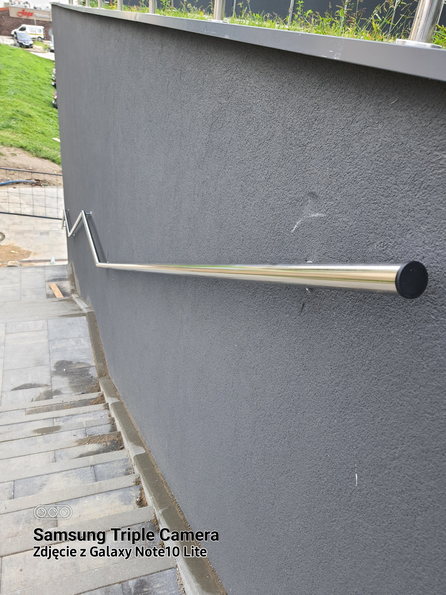 Zewnętrzna, nierdzewna poręcz schodowa zamontowana przy szarej ścianie z tynkiem strukturalnym, widok z góry na schody wyłożone szarą kostką brukową.