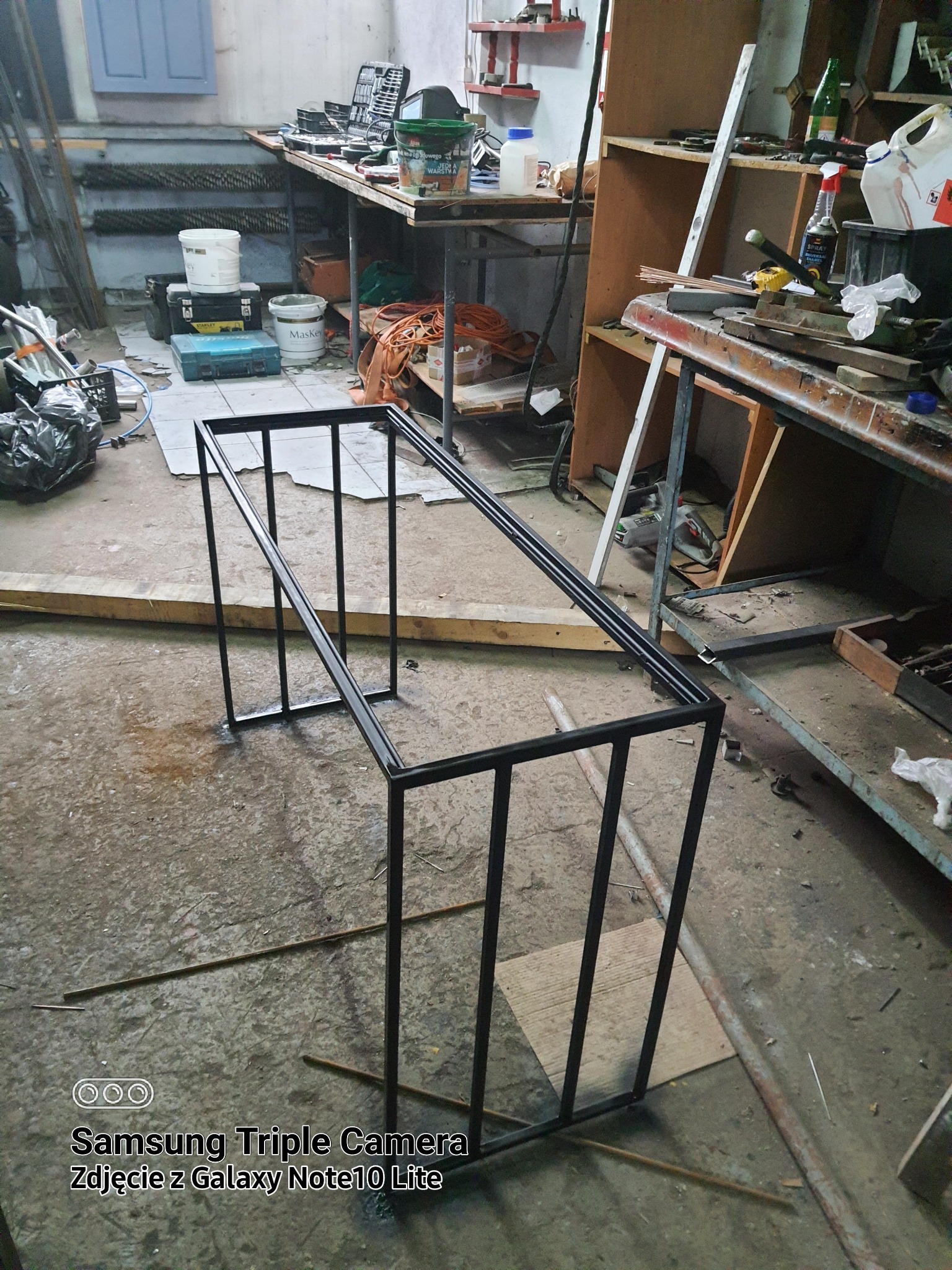 Surowa, czarna, metalowa rama stolika kawowego w warsztacie, widoczne ślady spawania, w tle narzędzia i materiały.