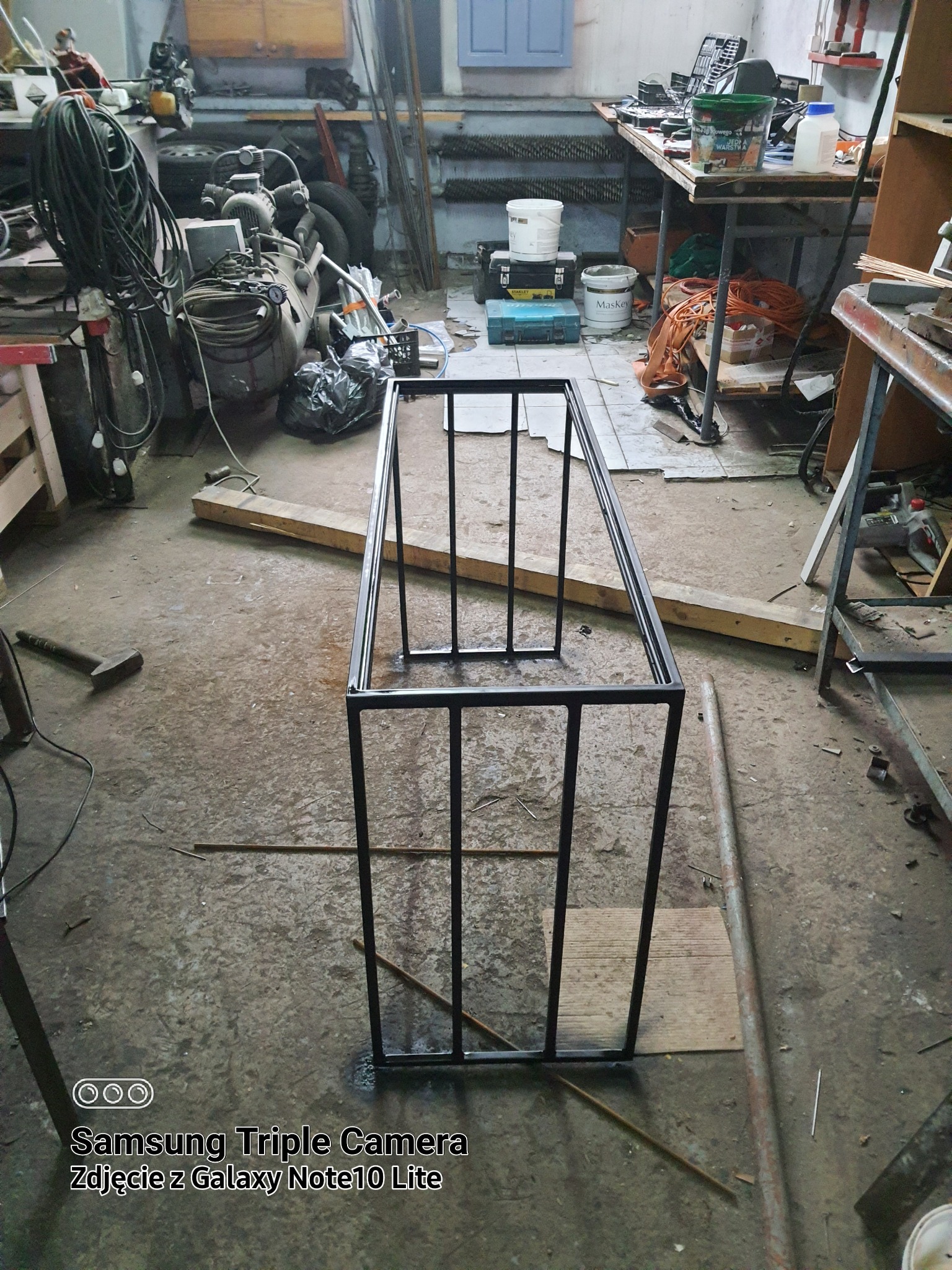 Surowa konstrukcja metalowa z czarnych profili w warsztacie, widoczne ślady spawania, narzędzia i materiały w tle.