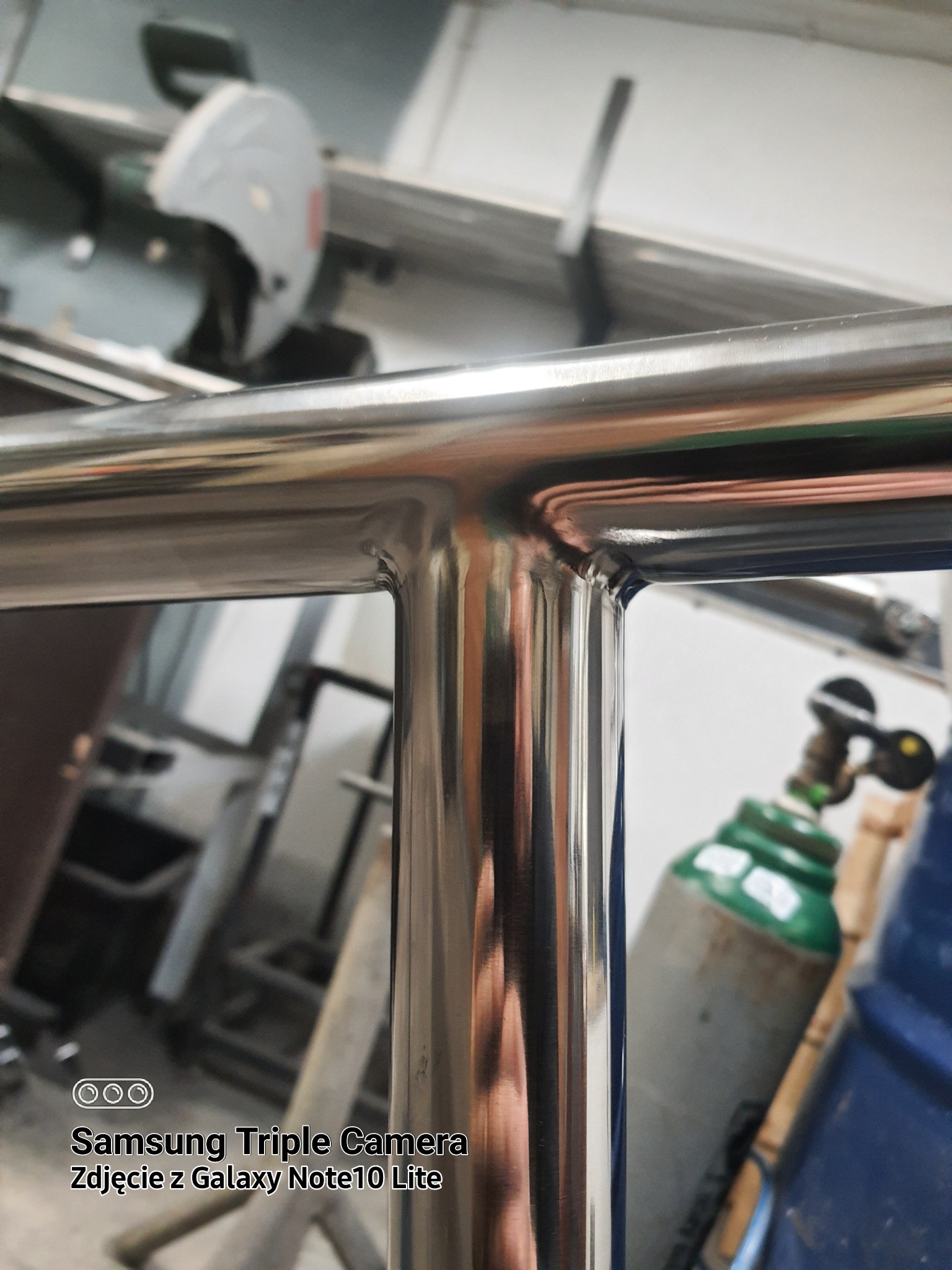 Szczegółowe zbliżenie na spawany, polerowany narożnik balustrady ze stali nierdzewnej, widoczne ślady spawania, w tle butla z gazem i piła do metalu.