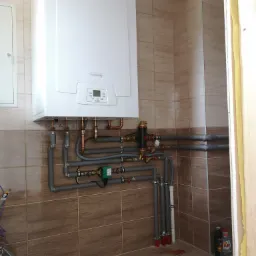 Biały wiszący piec gazowy marki De Dietrich zamontowany na ścianie z brązowych płytek, z widocznymi rurami miedzianymi i szarymi rurami instalacyjnymi oraz elementami armatury. W tle widoczny komin...