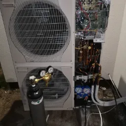 Otwarta obudowa dwu-wentylatorowej pompy ciepła z widocznymi elementami instalacji wewnętrznej, manometrami i butlą techniczną z gazem chłodniczym.