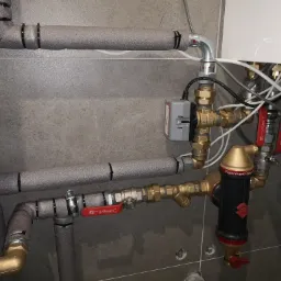 Szare rury grzewcze z izolacją termiczną, zawory z czerwonymi uchwytami i czarny filtr 'Flamcovent Clean Smart' zamontowane na ścianie z płytek ceramicznych w kotłowni.