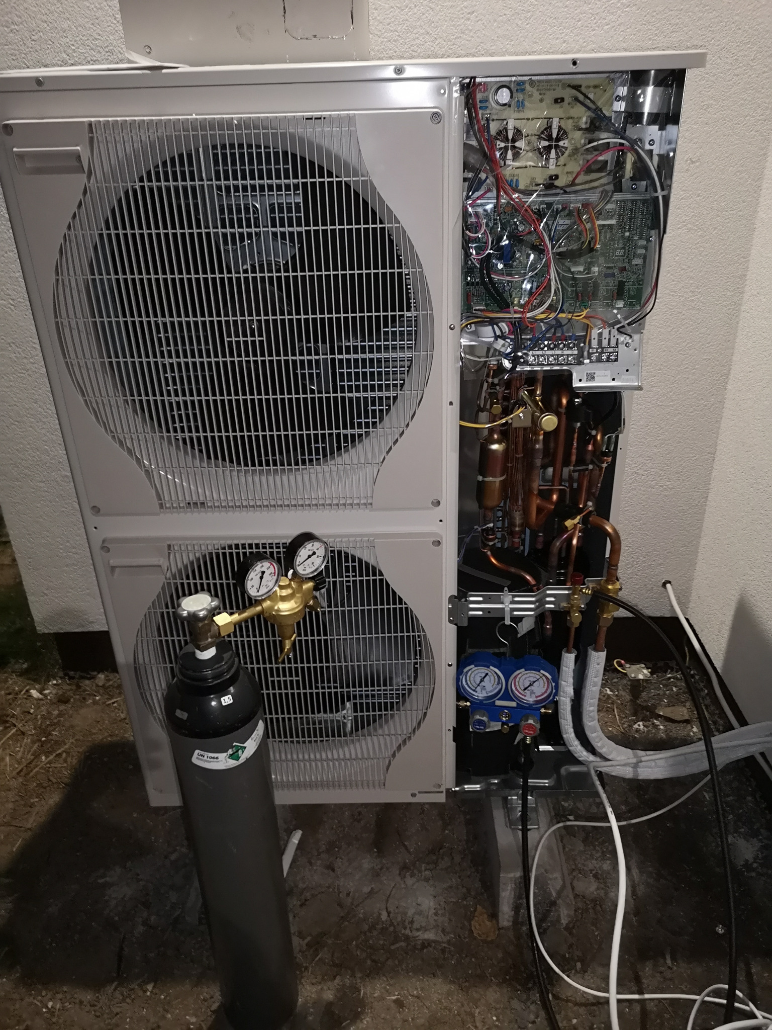 Otwarta obudowa dwu-wentylatorowej pompy ciepła z widocznymi elementami instalacji wewnętrznej, manometrami i butlą techniczną z gazem chłodniczym.