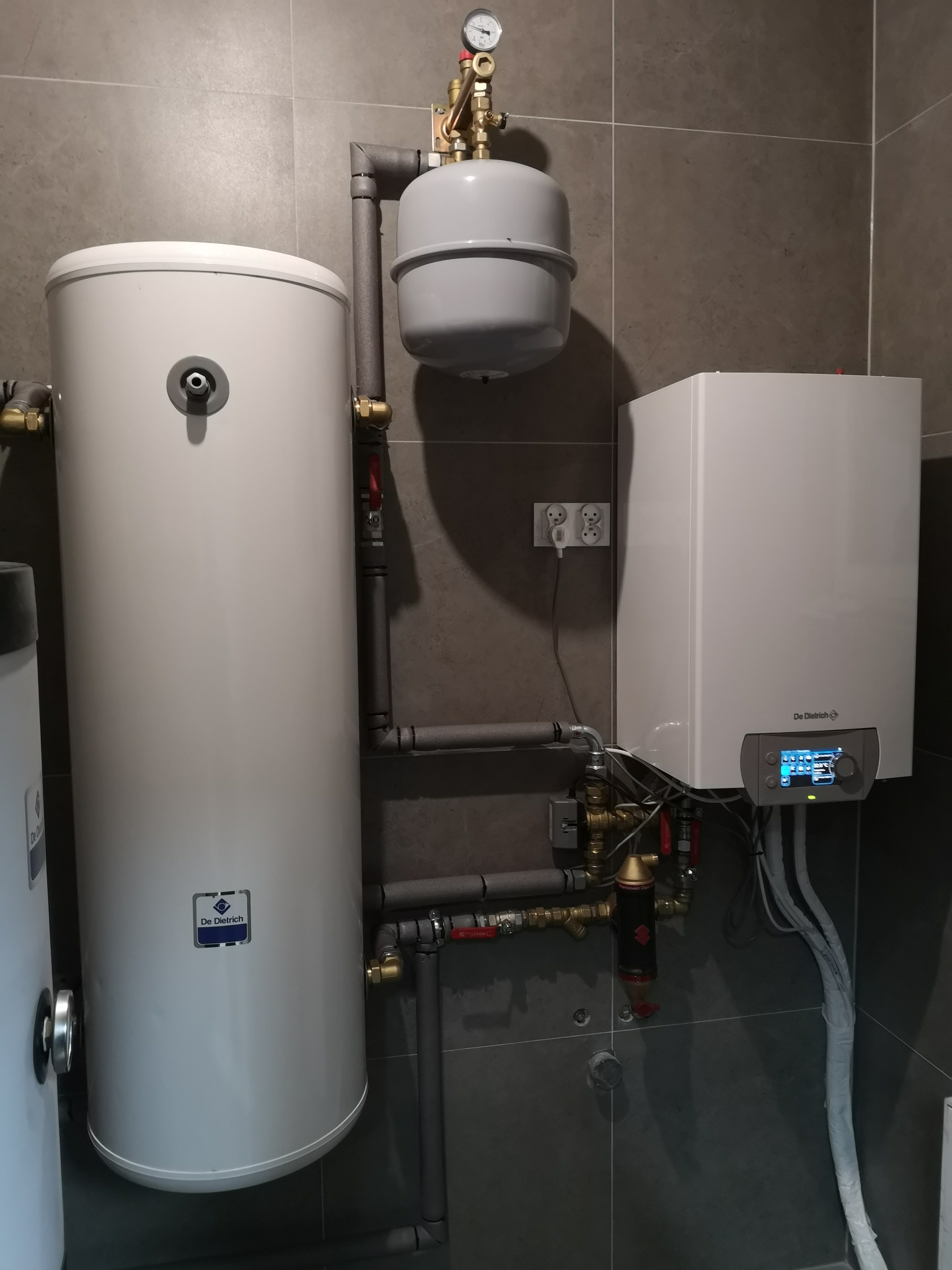 Nowoczesna kotłownia z białym zasobnikiem ciepłej wody i kotłem gazowym De Dietrich na tle szarych płytek, widoczne rury i armatura.