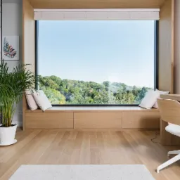 Jasna drewniana podłoga w pokoju z dużym oknem z widokiem na zieleń, biurko z krzesłem i doniczkowa roślina.